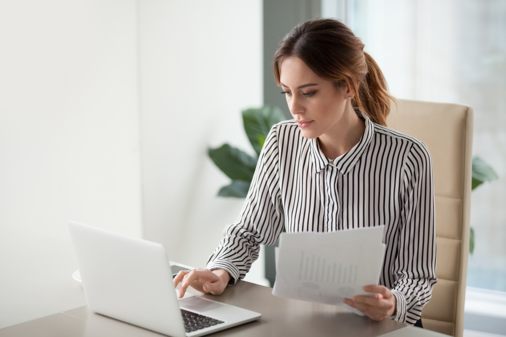 Serious focused businesswoman typing on laptop holding papers preparing report