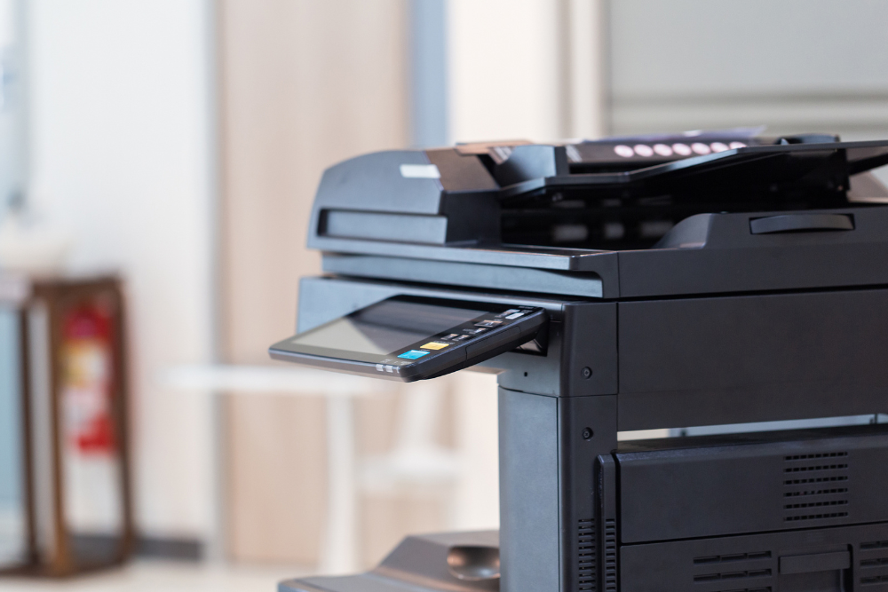 A close-up of an office copier in the foreground with a blurred-out office setting in the background.