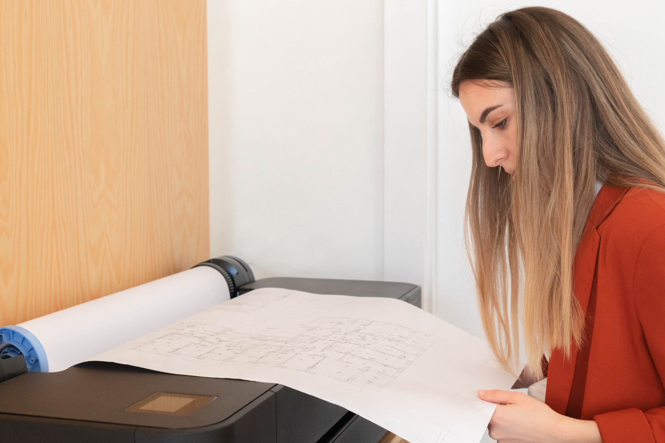 A woman reviews a large printed sheet of architectural or construction drawings as it is printed out from a wide format printer.