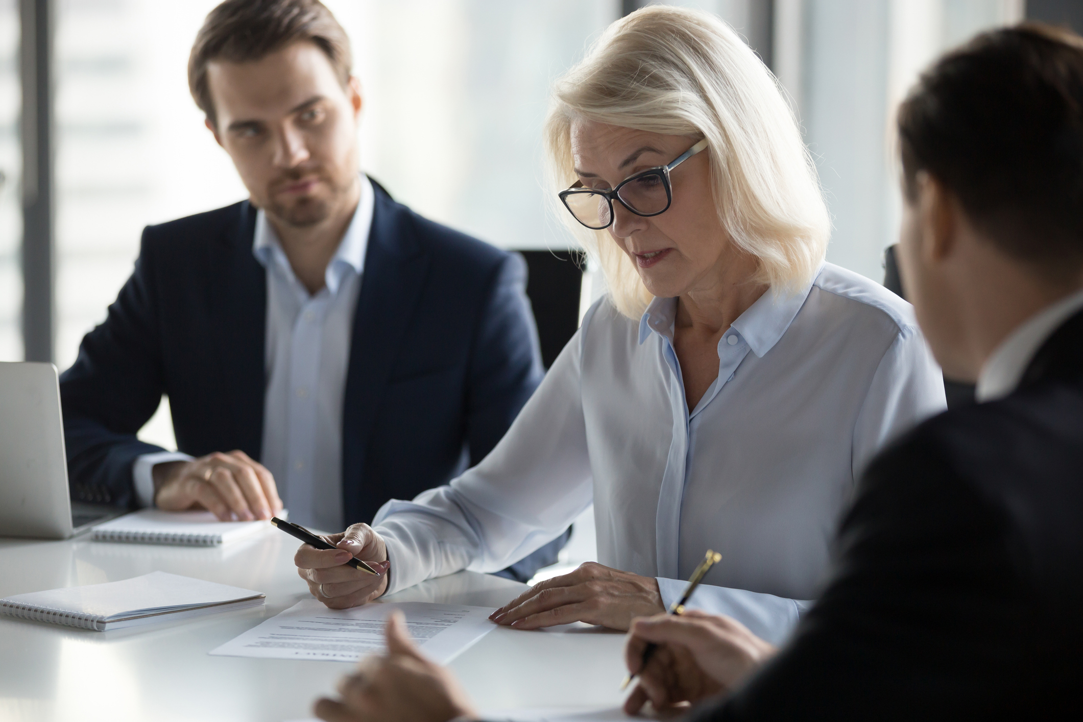 Businessmen sitting at desk headed by middle aged serious concentrated female in eyeglasses checking agreement document before signing it. Financial director ready affirm official paper with signature signifying copier leasing companies.