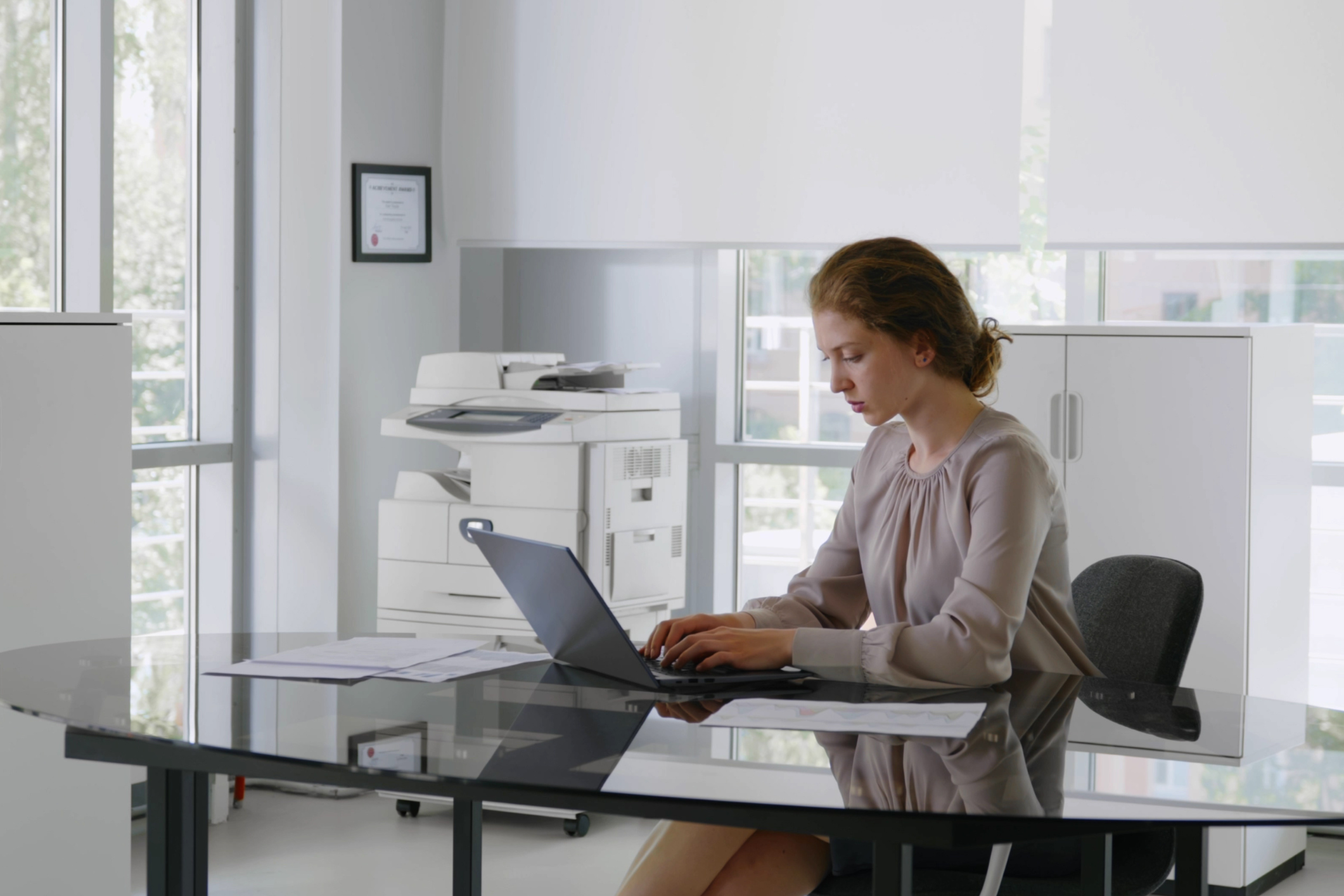 A businesswoman sits at a desk in a clean, bright, modern office with an office copier in the background.