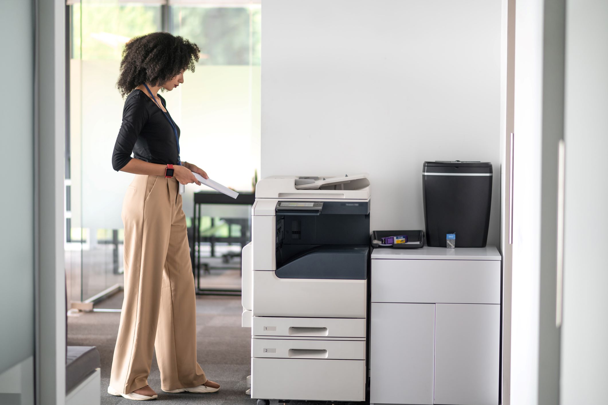 A stylish professional stands over an office printer and scanner waiting for outputs.
