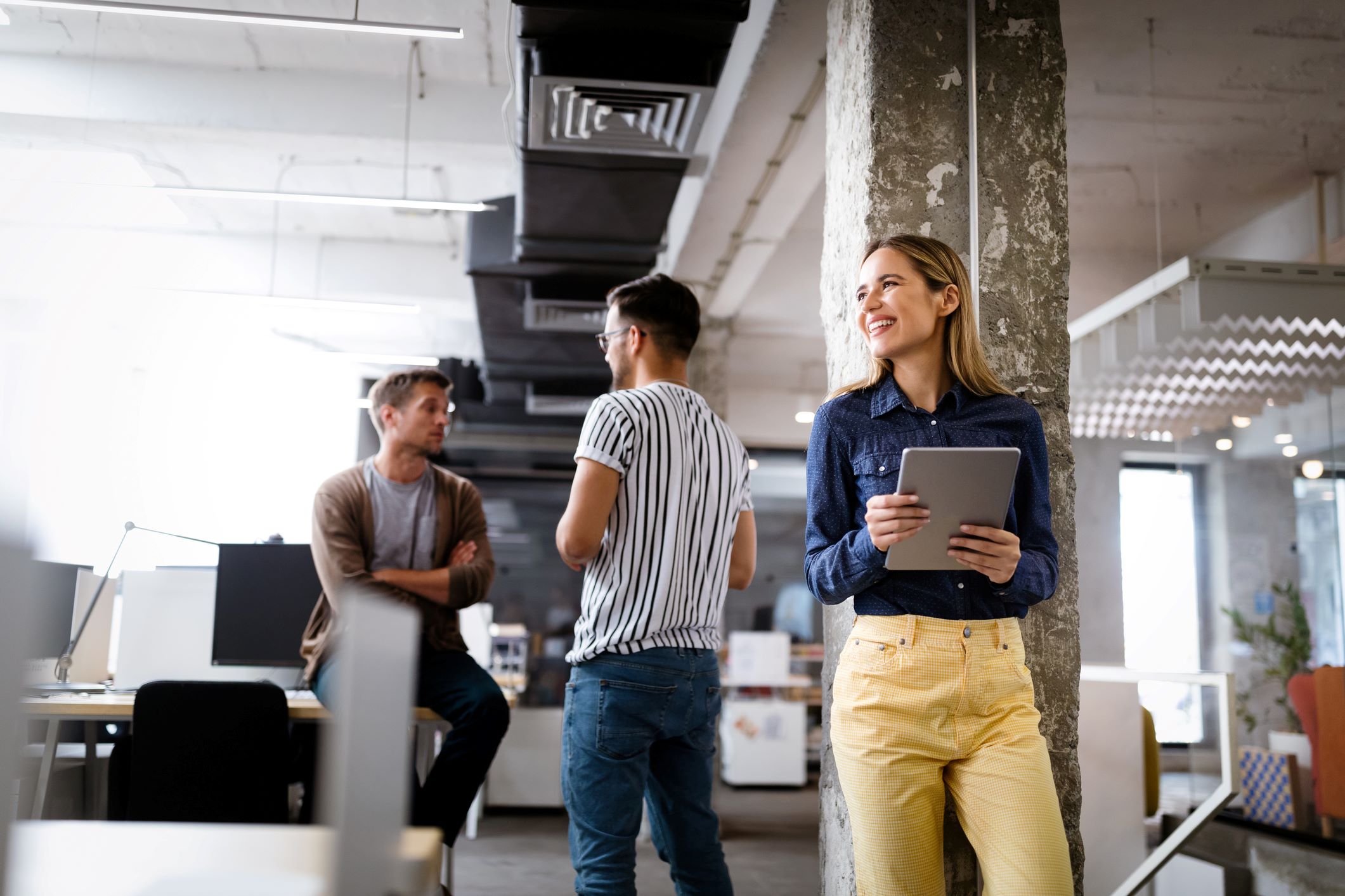 A smiling professional stands in the foreground of a modern office while two coworkers chat in the background symbolizing a printer rental.