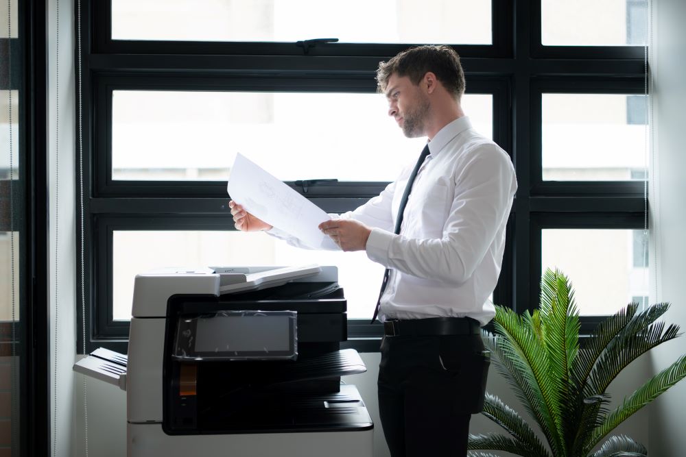A professional stands in front of a copier examining the output with a questioning expression, signifying the need for copier service.