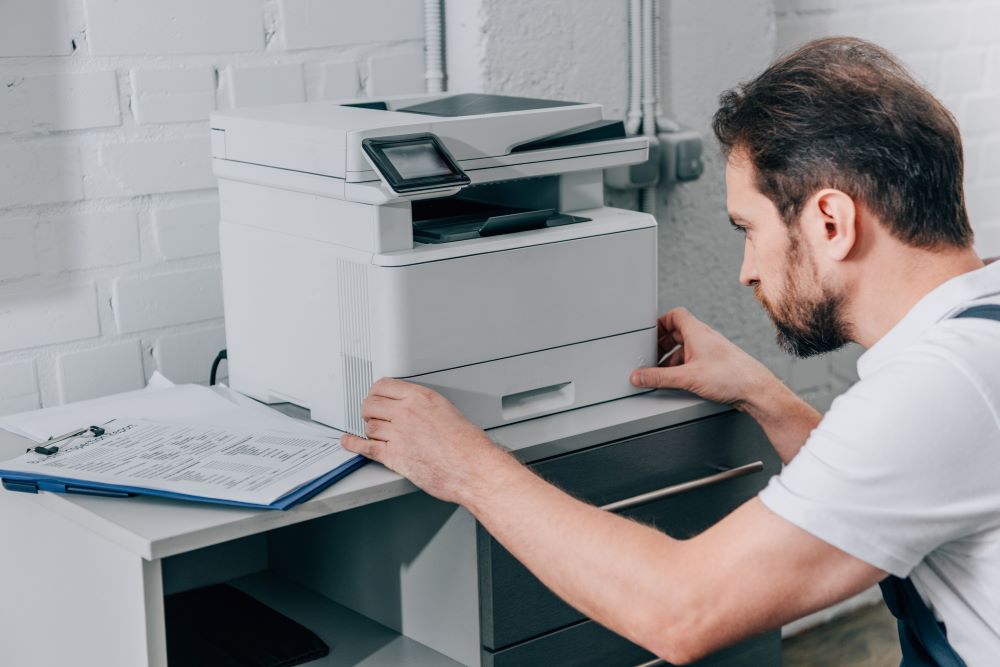 A repair service technician examines a copier on a table in an office with a checklist on a clipboard next to him signifying copier service provider.