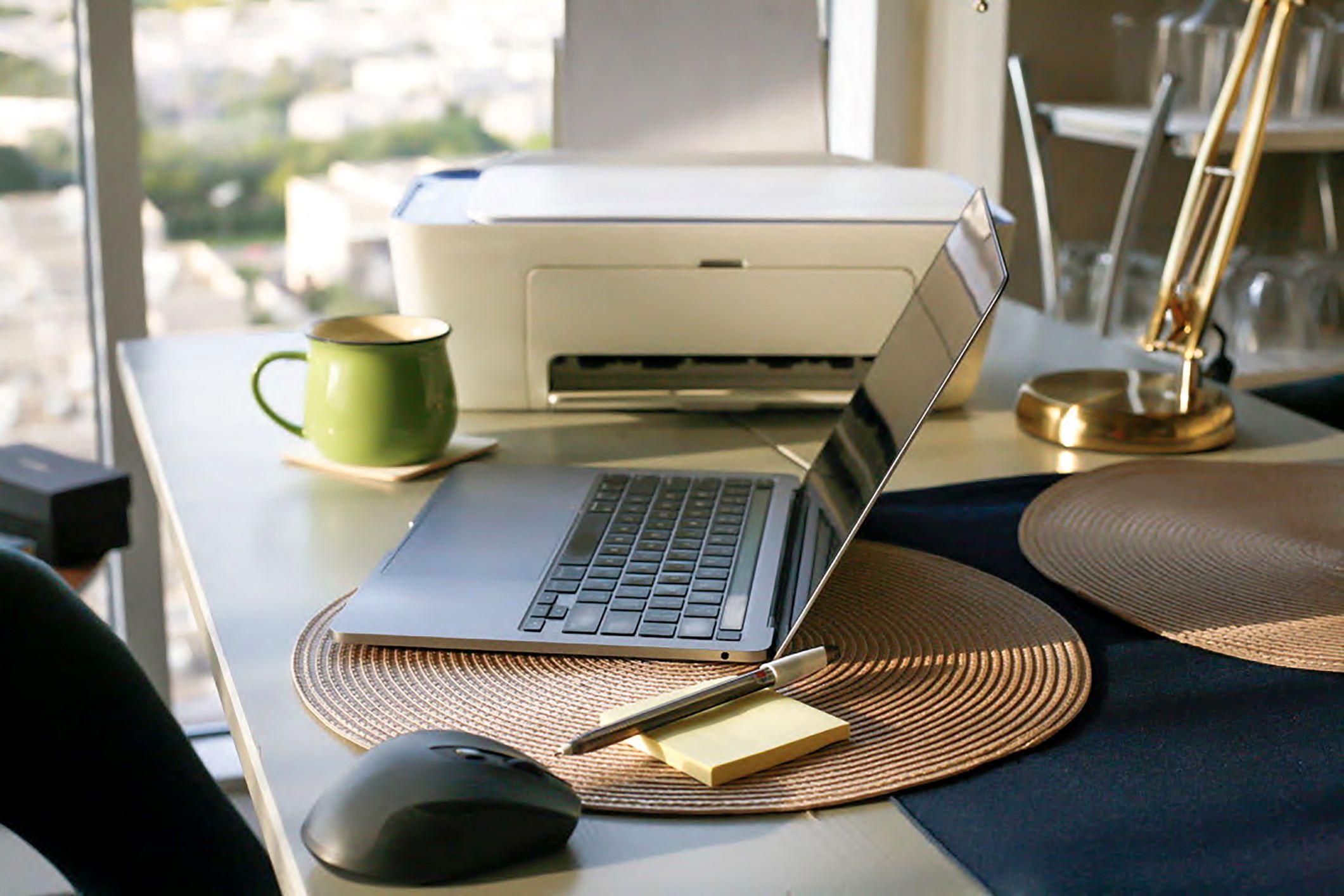 An office setup featuring a sleek laptop on a placemat, a business printer, a green mug, a wireless mouse, sticky notes with a pen, and a white printer on a desk with a city view from a window.