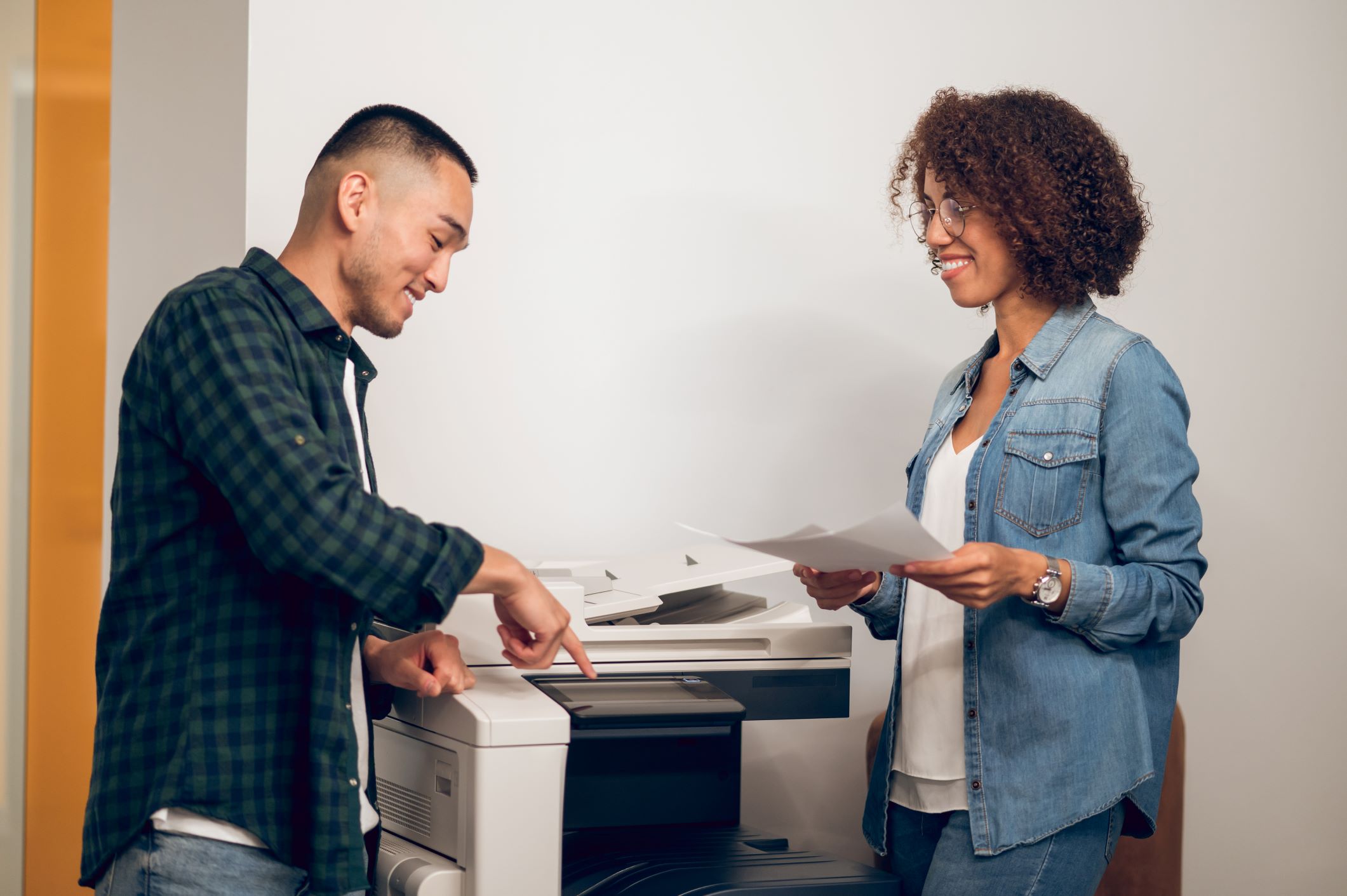 A smiling man and woman interacting in an office setting, with the man using an office copier while the woman reviews a printed document.