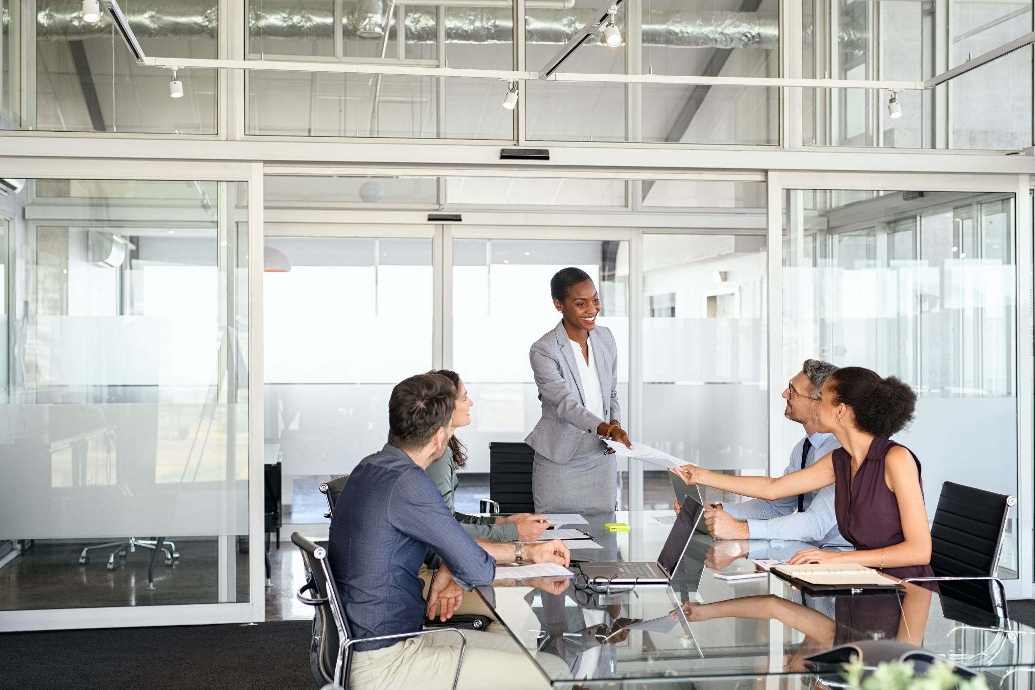 The image showcases a bright and modern office meeting room where a business transaction is taking place. A well-dressed woman stands handing a document to a seated man across a glass table, with another woman seated beside him. The trio appears engaged in a professional interaction, possibly discussing terms or finalizing an agreement such as a copier lease. Their expressions are friendly and cooperative, indicating a positive negotiation or consultation. The room is stylish and contemporary, with glass walls that allow natural light to flood in, and the office's interior design suggests a corporate setting. The exchange of documents is central to the scene, highlighting the importance of paperwork in official business arrangements, including leasing office equipment like copiers.