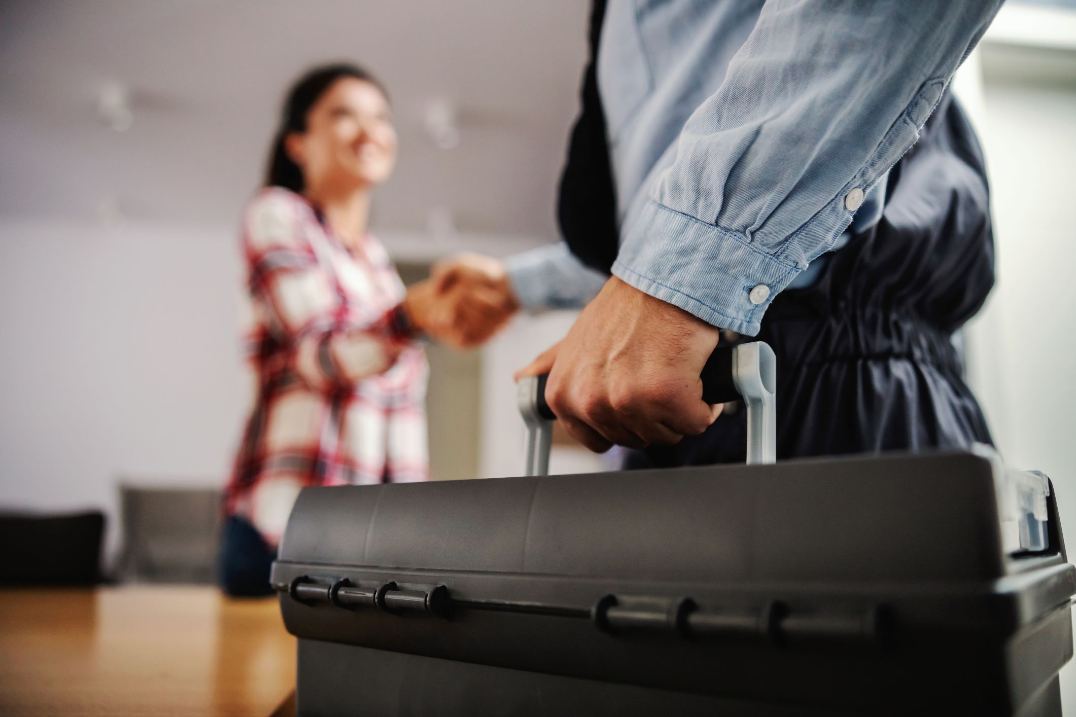 The image depicts a close-up view of a person's hand gripping the handle of a large, portable tool case, with the focus on the case suggesting that they are a technician arriving for a job. The case is sturdy and professional, hinting at technical equipment inside, possibly for copier service or maintenance. In the softly blurred background, a smiling woman is seen, likely greeting the technician, indicative of a positive customer service experience. This scene is representative of professional copier service technicians who travel to businesses to perform repairs, maintenance, or servicing on office copiers and printers, ensuring that office equipment functions smoothly and efficiently.