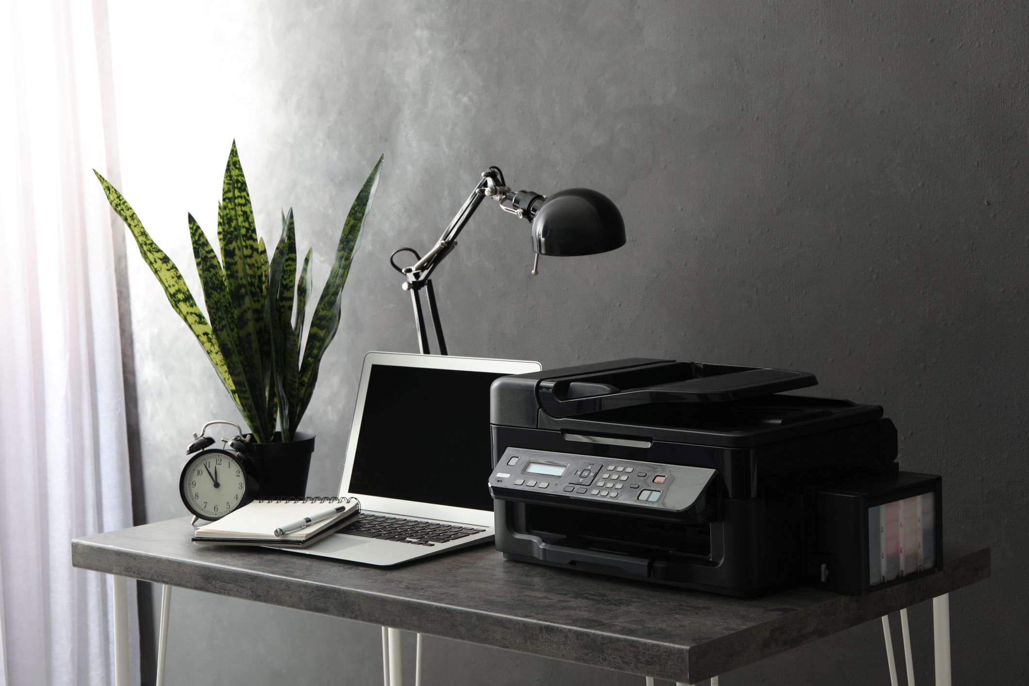 The image presents a neatly organized workspace, featuring an office printer as the central element. The printer is a modern, black multi-function device, capable of printing, scanning, and copying. To its left, there's a laptop open on a desk, alongside a spiral notebook and a silver pen, ready for note-taking. A classic black analog clock indicates the time, while a potted plant with long, variegated leaves adds a touch of greenery and life to the scene. A desk lamp with an adjustable arm is poised above, providing focused illumination. The overall setting suggests a professional and efficient environment, with the office printer serving as an essential tool for daily business operations and document handling.