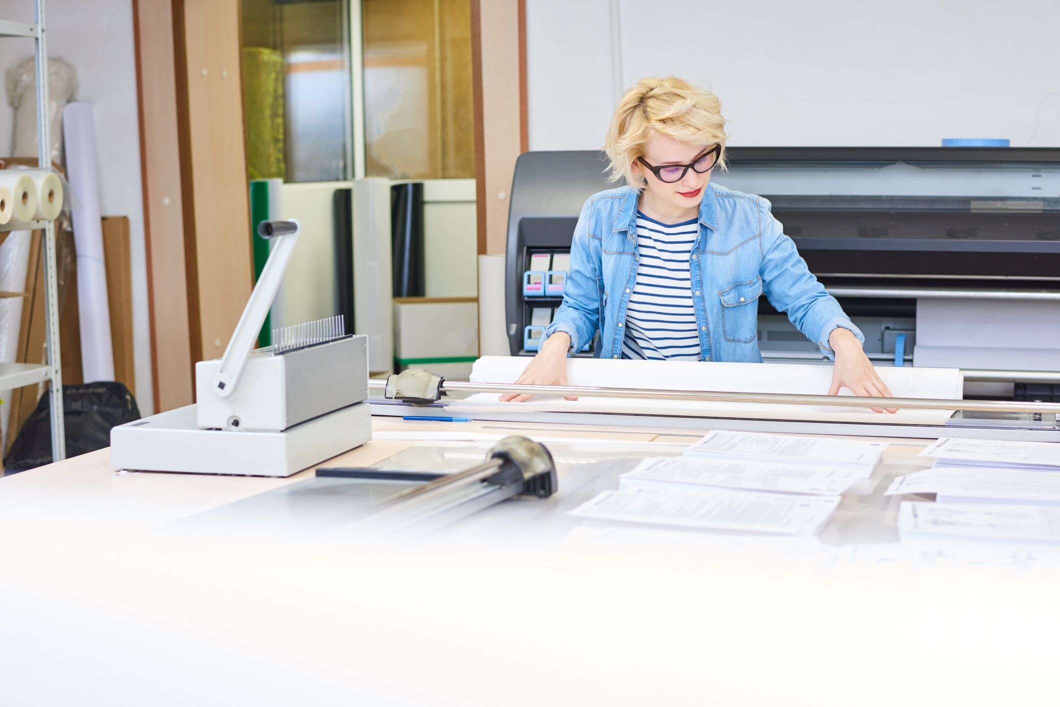 A focused woman with blonde hair and wearing glasses is working in a print shop, standing behind a wide format printer. She is aligning a large sheet of paper on the cutter in a bright, busy office environment, with various printing supplies and equipment around her.