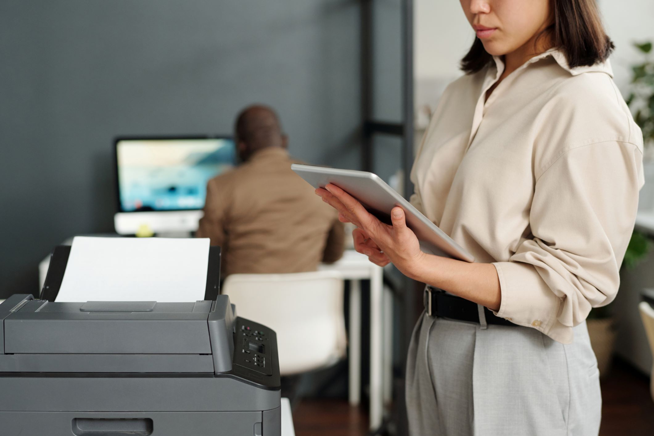 A professional setting where a woman is focused on her tablet next to an all-in-one office printer, with a male colleague working on a computer in the blurred background, highlighting the multifunctionality and workplace integration of such a device.
