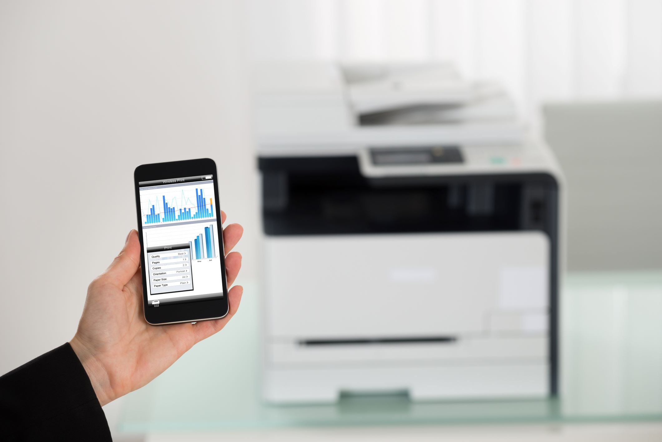 A hand holding a smartphone with a graph on the screen, symbolizing data analysis, with a large multifunctional office printer in the blurred background, representing the convenience of mobile printing technology in a modern office setting.