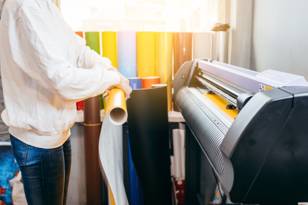 A person operating a plotter printer in a brightly lit workspace, surrounded by rolls of colorful printing materials.