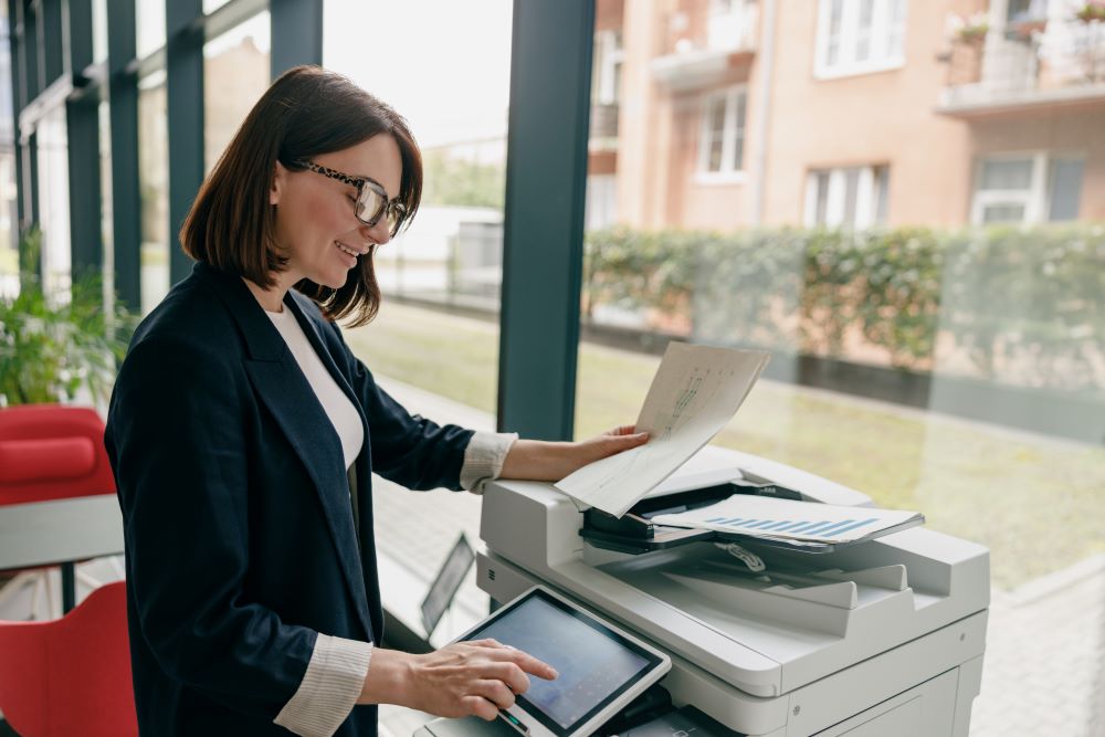 Smiling businesswoman using an Office MFP to scan or copy documents in a bright office setting.