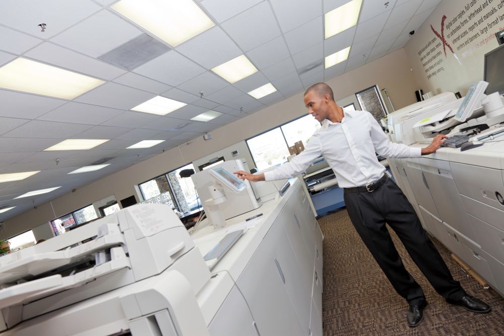 A well-dressed businessman stands in front of a production printer interacting with its interface.