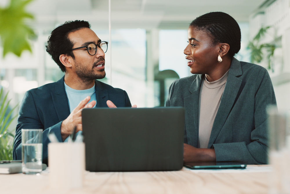 Two modern employees sitting at a desk having a discussion behind a laptop signifying lease a business copier