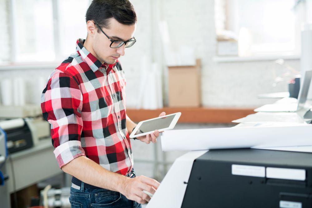A young employee uses a tablet at a large format printer signifying mobile device printing