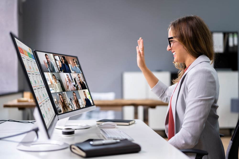 A smiling young professional sits at a desk behind two screens, where you can see the video chat with nine additional professionals waving hello to signify the importance of unified communications.